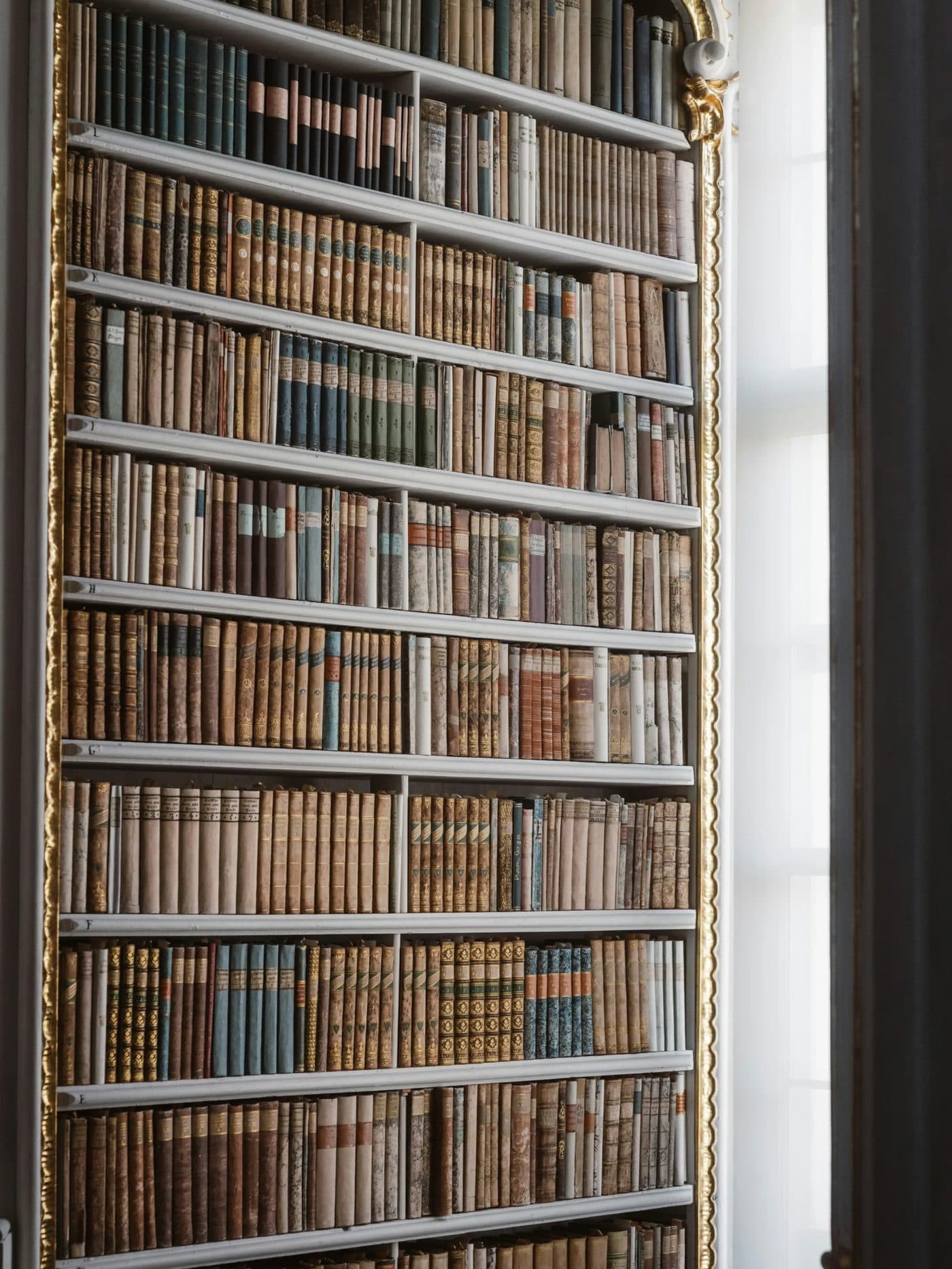 Tall gilded bookcase with ordered rows of leather and cloth-bound volumes beside a curtained window.