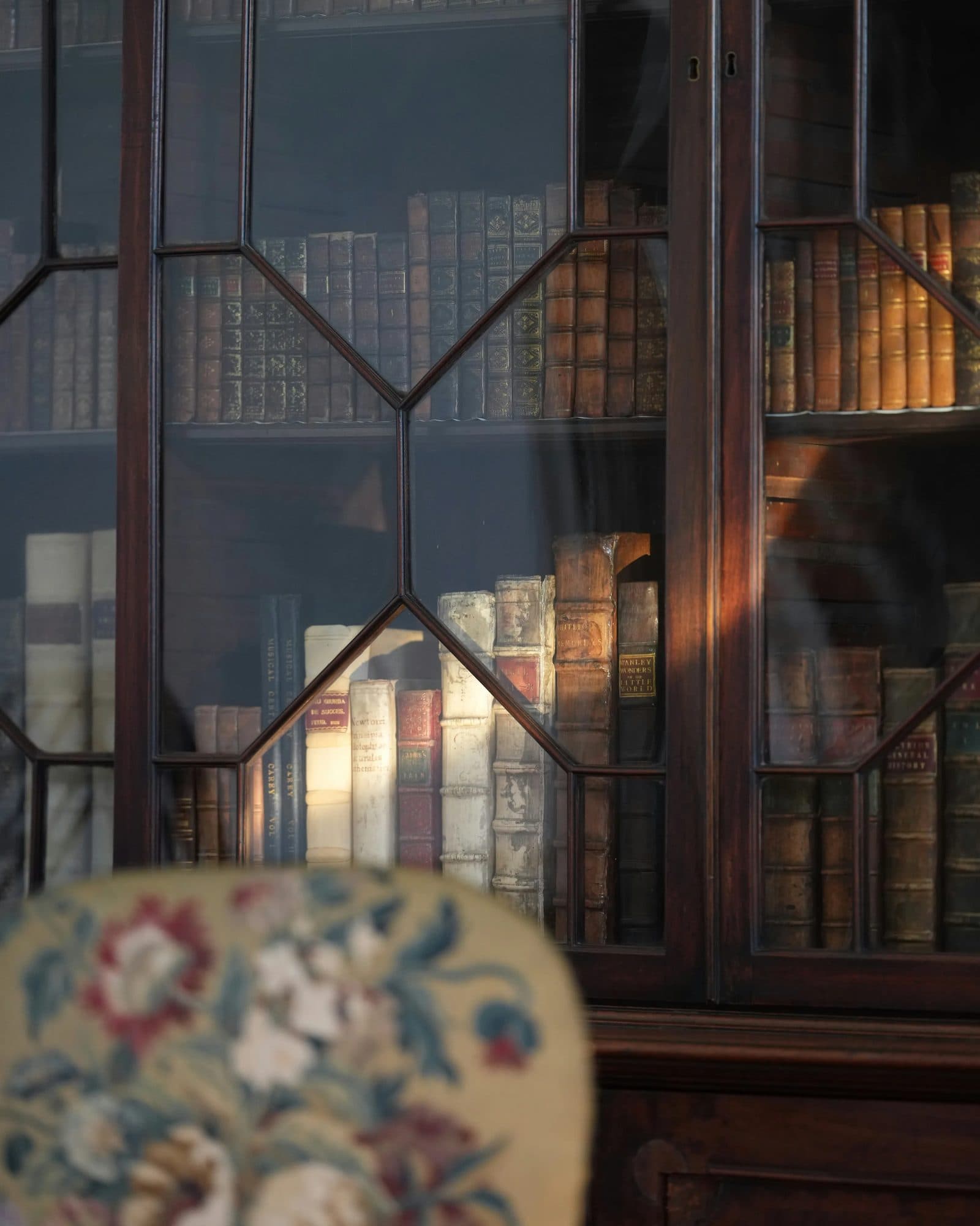 A period mahogany bookcase with glass doors beside an upholstered chair in warm natural light.